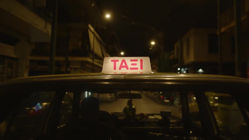 Male cabbie drives a taxicab down a street lined with parked cars at night