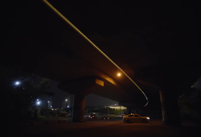 A yellow taxi cab sits on a dark street under an elevated roadway illuminated by artificial lights