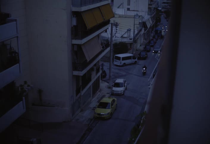 Looking down from an elevated position a dimly lit urban street features parked cars along the sides of tall buildings as dusk settles