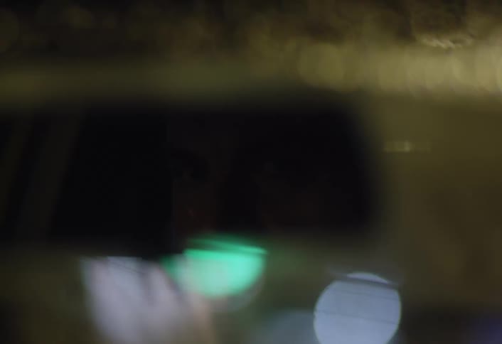 A close up of a rearview mirror on a car windshield shows a city at night with rain and wipers in motion
