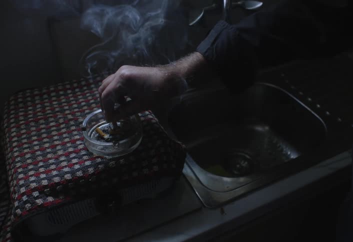 A hand drops a cigarette butt into a glass ashtray filled with other butts with smoke rising in a dimly lit space near a sink