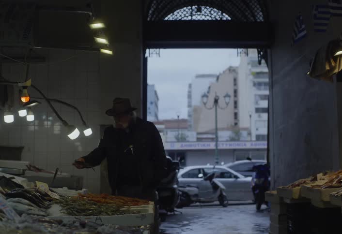 A person wearing a hat examines produce displayed under artificial lights in a market with a view of a city street outside