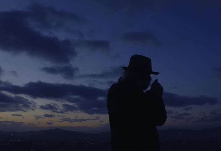 A silhouetted figure in a hat and coat stands under a twilight sky lighting a cigarette