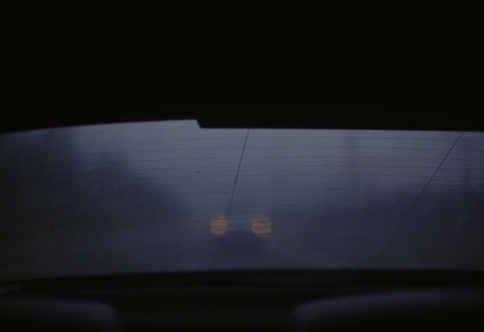 A misty car windshield reveals a vehicle behind with flashing headlights in the twilight darkness