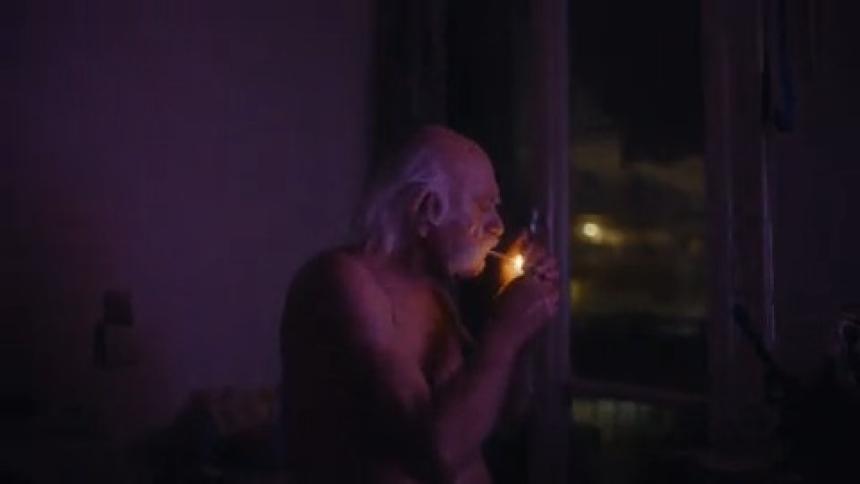An elderly man with a mustache and white hair lights a cigarette while switching channels on a tv in a room