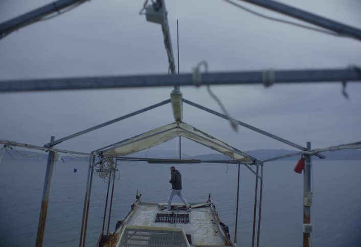A lone figure walks on the deck of a boat under a gray overcast sky and above a calm body of water