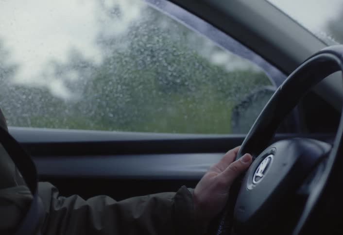 A close up shot from inside a car shows a persons hands on a steering wheel as they drive in the rain