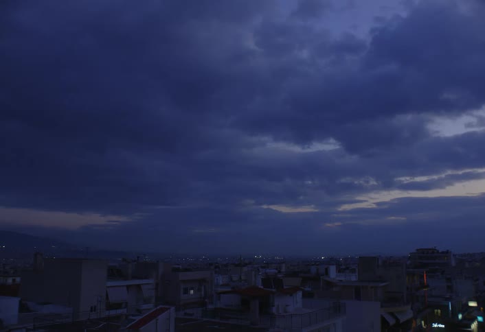 A cityscape is viewed under a dark cloudy evening sky with hints of light on the horizon