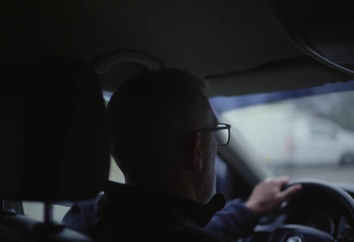 White haired man steers his car along a road passing parked vans