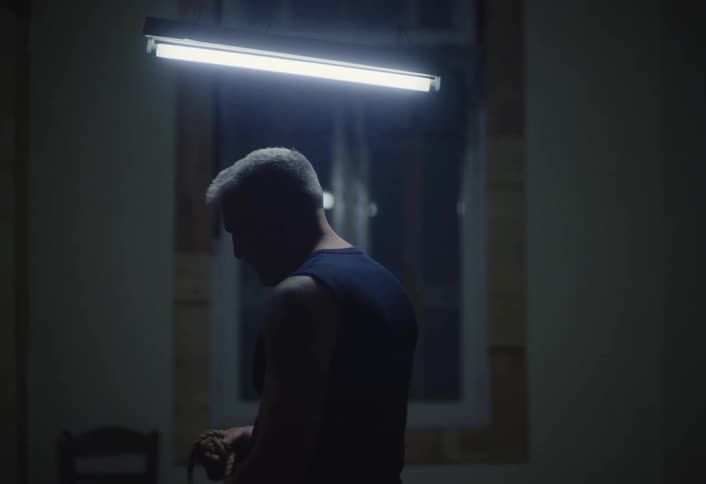 A person with short hair stands in a dimly lit room under a bright fluorescent light fixture handling a rope