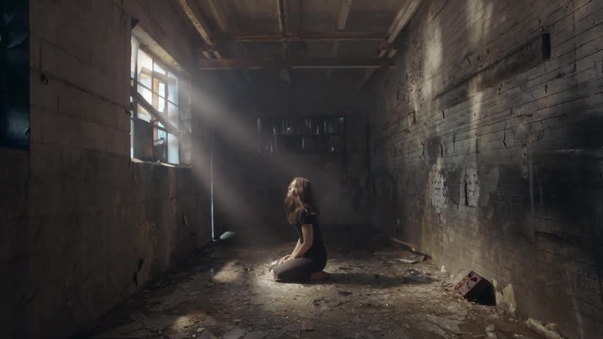 Woman kneels in a dirty building in front of a window as sun rays drench her