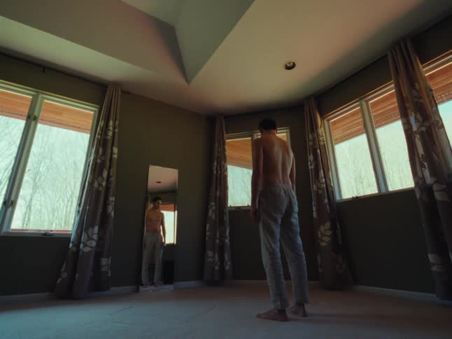 A shirtless man stands in a room adorned with numerous windows and curtains gazing thoughtfully at his reflection in a mirror