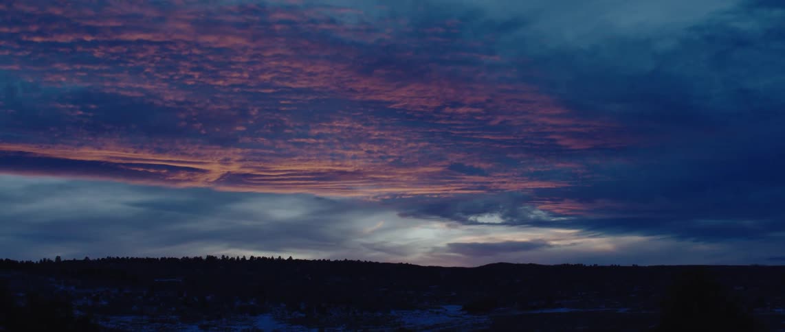 A dramatic sunset sky is painted with streaks of orange and pink hues amidst dark moody clouds casting a dim light over a silhouetted landscape below