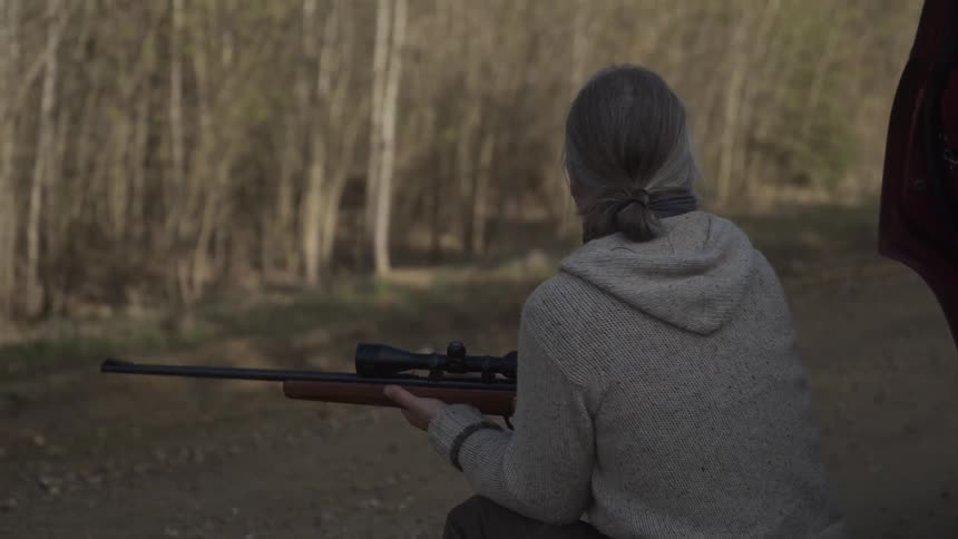 Older man takes a shot with a rifle while half kneeling near his parked vehicle