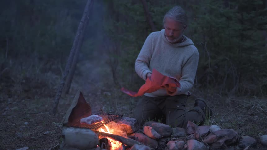 Man placing steaks on grill in an open campfire in the wilderness