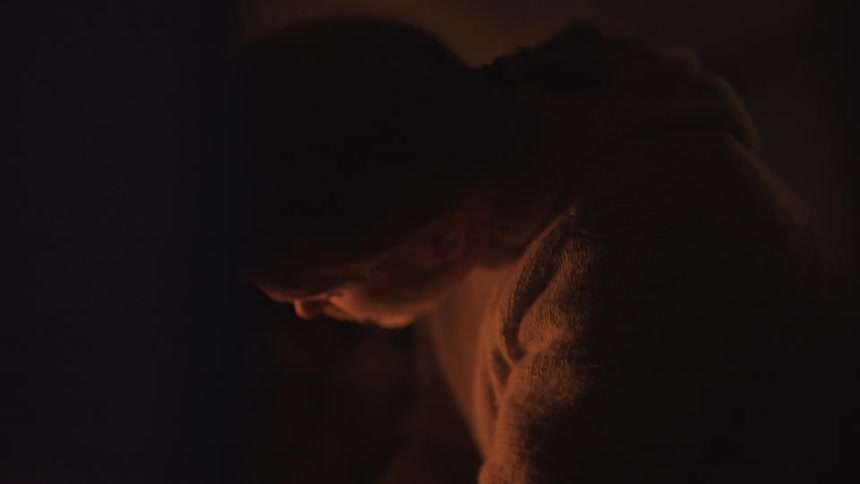 Warm light from the left illuminates the profile of a hooded person looking down in a dark room