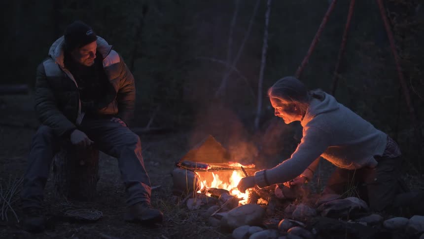 Two men are cooking food over a campfire in a dark forest setting