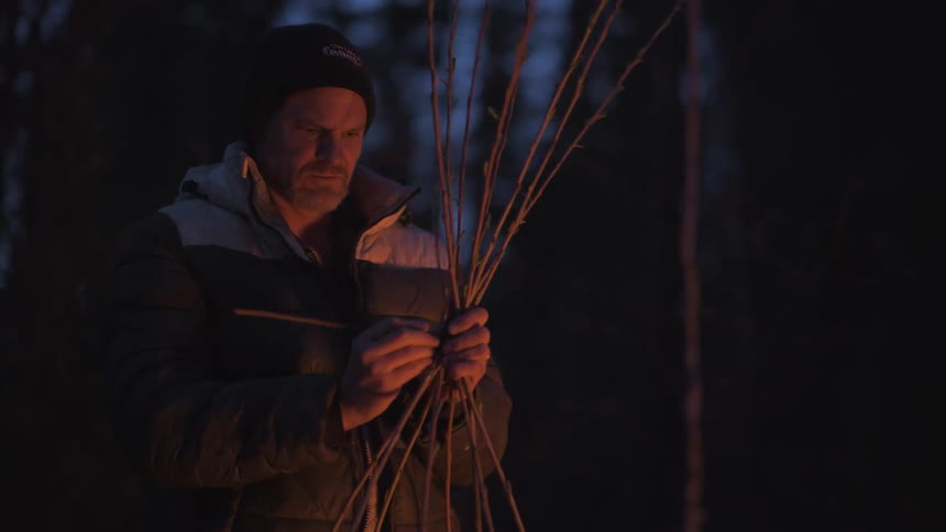 Man weaves small branches together while lit by the light from a campfire