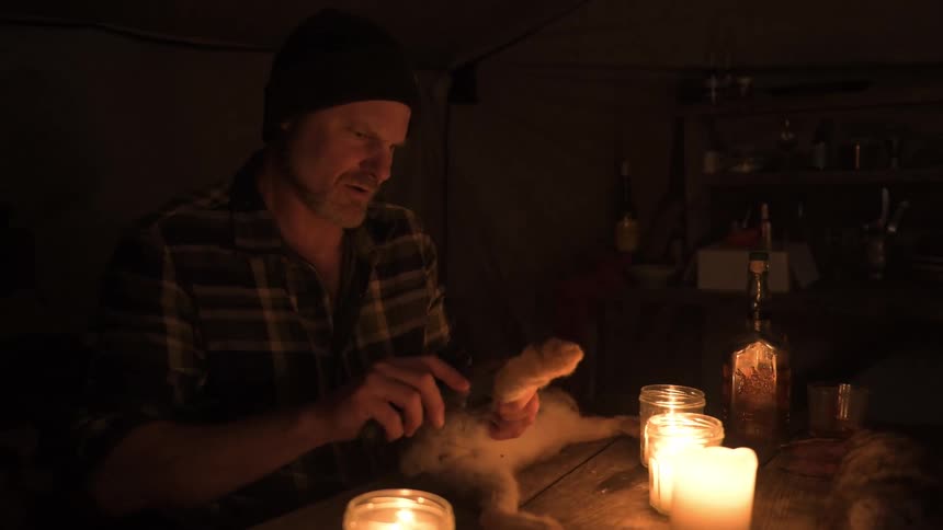 A hunter sits in a dimly lit room at night using a knife to skin a rabbit on a candlelit table