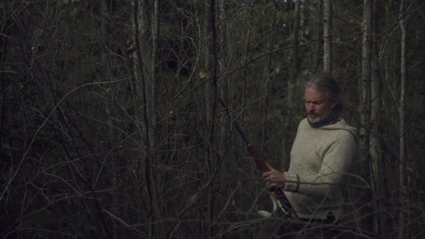 A man with long gray hair wearing a light colored sweater and carrying a rifle and hunted rabbits walks through a dense dark forest