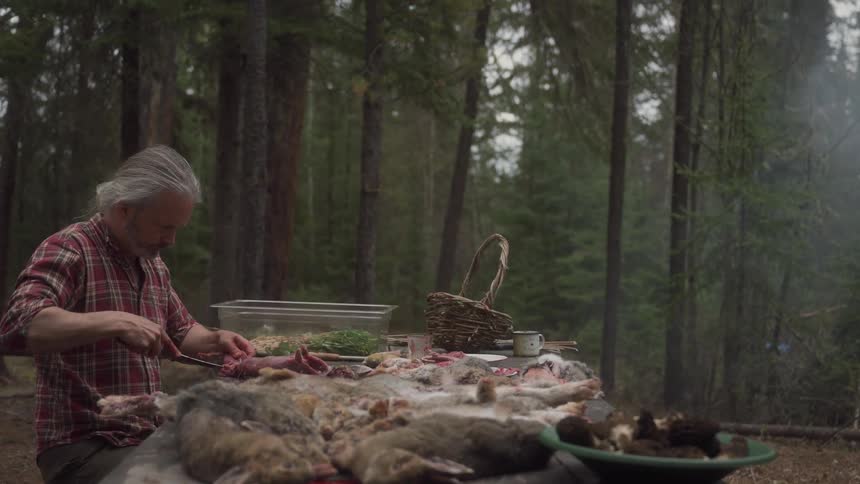 A hunter in a plaid shirt processes animal carcasses on a table in a forest with smoke rising in the background