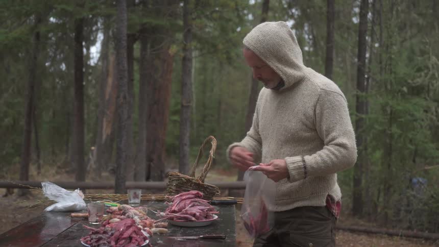 A man in a hooded sweater stands at a table in a forest handling pieces of raw meat