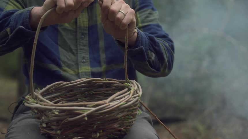 A man sits in a forest near a smoking campfire carefully handcrafting a wicker basket