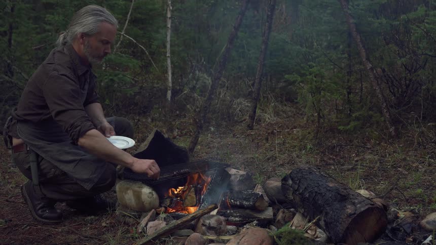 A man kneeling next to a campfire he appears to be picking up a skewer of meat off of the grate over an open flame he is in a camping setting out in a forest area and he appears to be getting ready to have his meal
