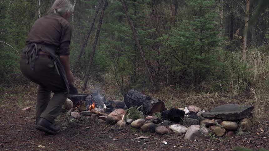 A man with gray hair kneels beside a stone lined campfire in a forest tending to the small flames beneath a makeshift tripod setup