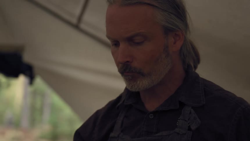 A closeup of a man with white hair tied up drinking from a glass inside a large tent in a forest