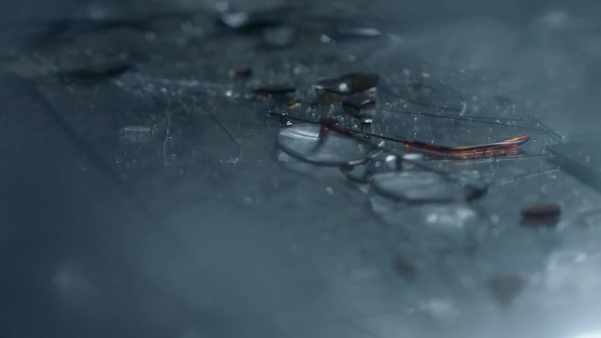 A pair of broken eye glasses rest on a floor strewn with broken glass