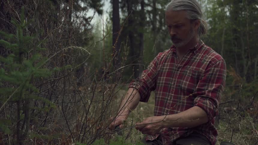A white haired man cuts small twigs using a knife in a forest clearing