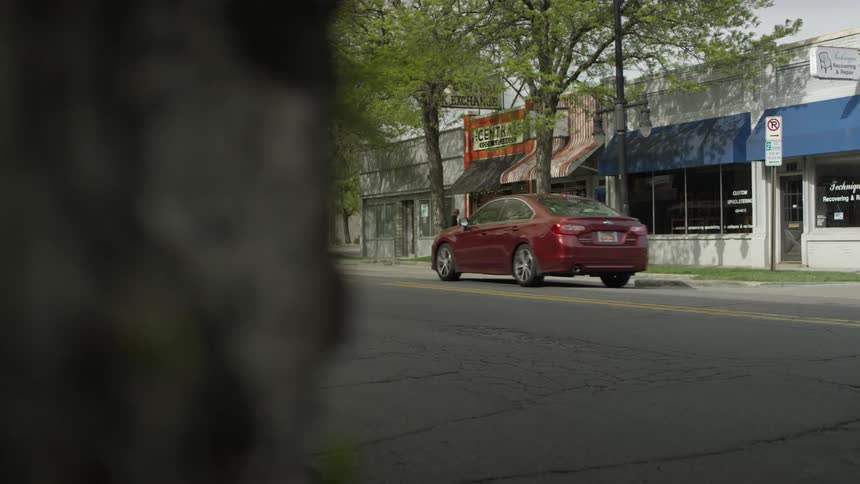 A car passes storefronts on a tree lined street