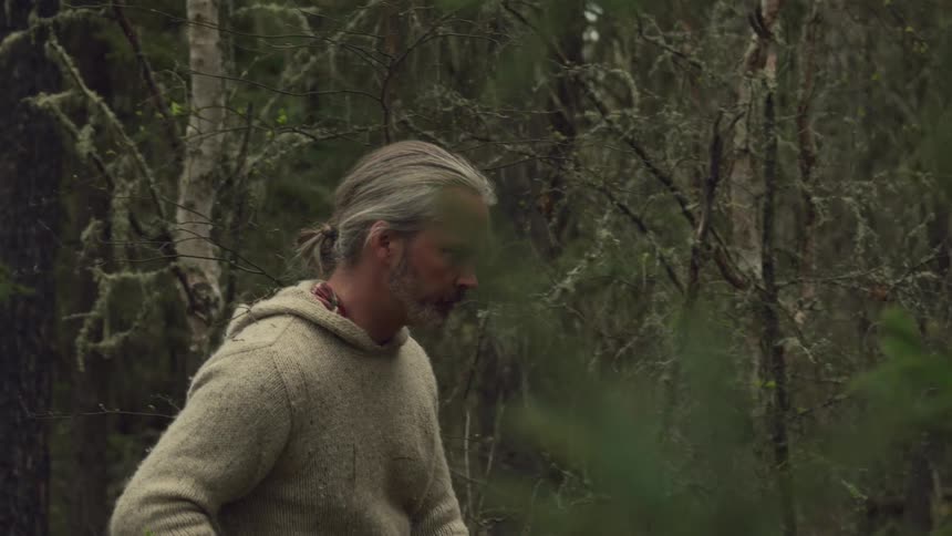 A man with long grey hair and a rifle walks through a dense forest during the daytime