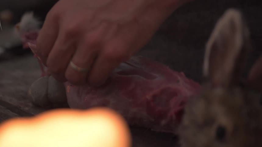 Man cuts a strip of meat from a carcass while butchering a dead rabbit outdoors during the day