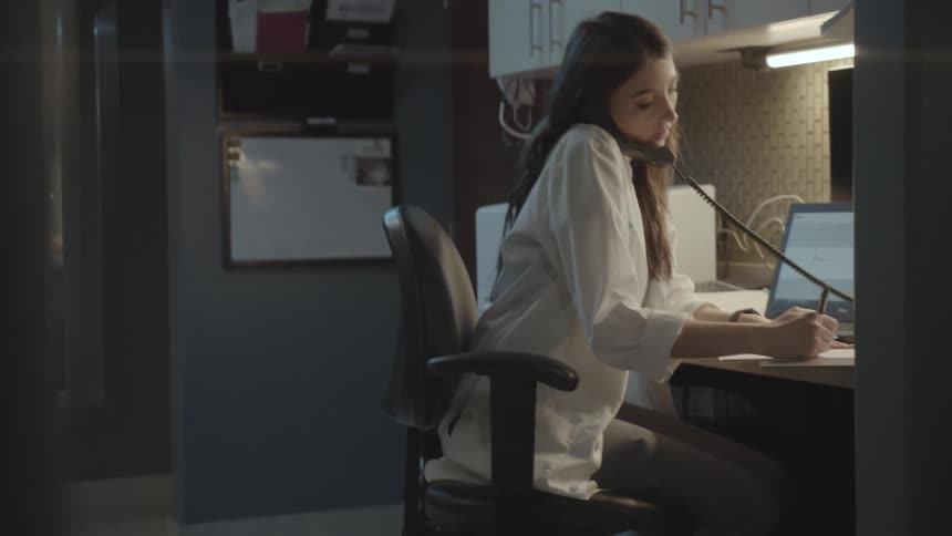 A woman sits in a cubicle engaged in a phone call while taking notes