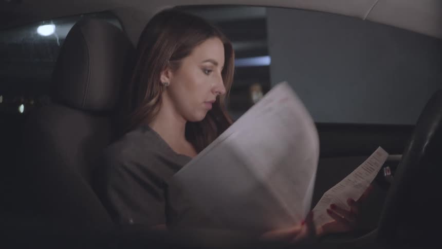 A woman with long brown hair sits in the drivers seat of a car at night illuminated by the interior light and reads through papers