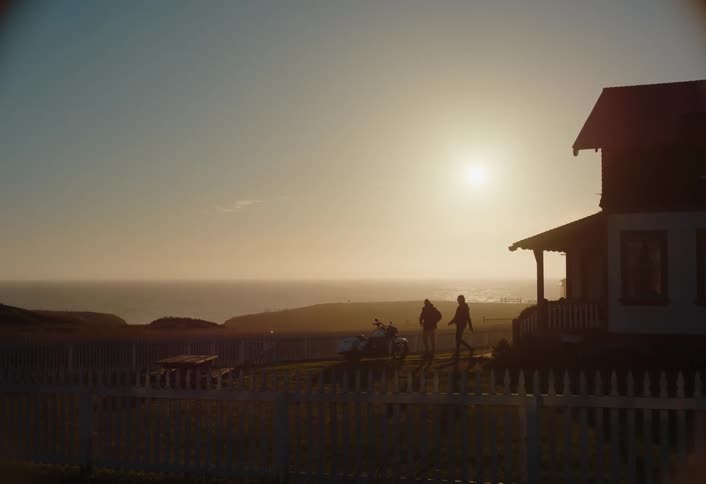 A couple walks out of a home with a picket fence passing a picnic table and a parked motorcycle as the ocean stretches to the hazy horizon under a setting sun
