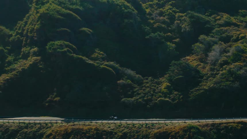 A motorcycle travels along a winding coastal highway nestled between lush green mountains and the ocean