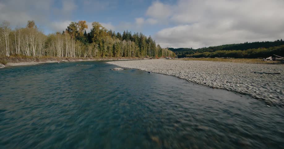 A wide flowing river with clear blue water stretches through a valley flanked by gravel banks and trees under a sky with scattered clouds