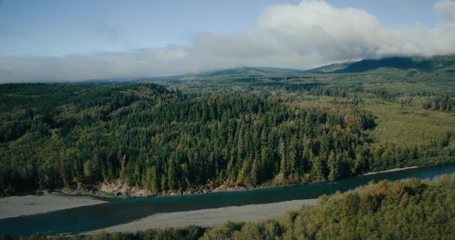 An aerial flyover captures a vast forested valley with a winding river flowing through lush green hills under a sky of scattered clouds