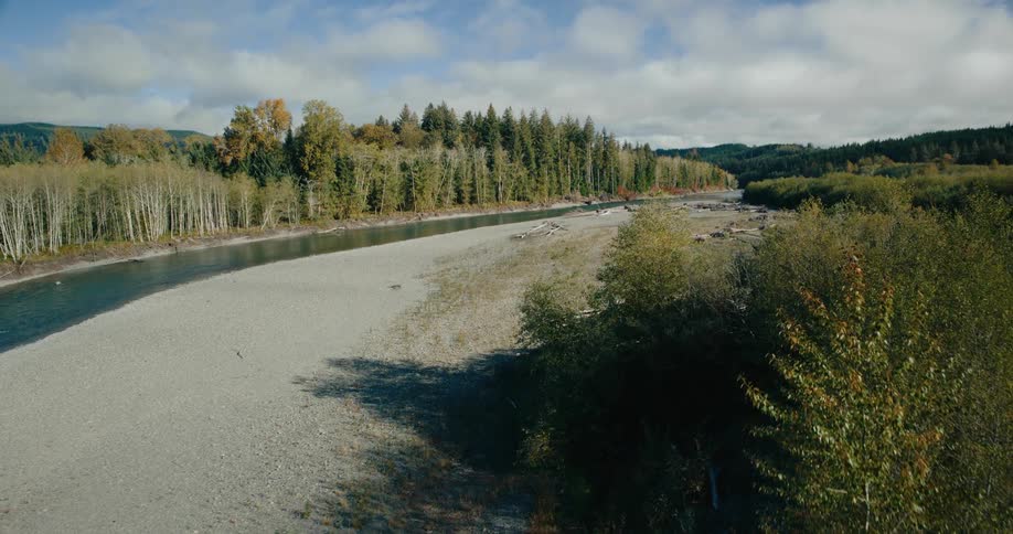 An aerial shot captures a wide river flowing through a valley with a gravel bar running alongside it surrounded by trees and vegetation under a partially cloudy sky