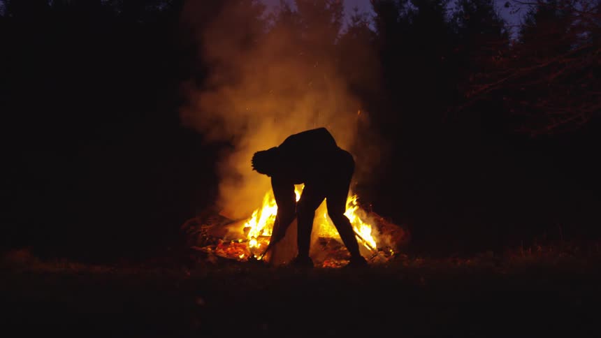 A man with a spade shovel is digging a perimeter line around a blazing bonfire sending smoke into the night sky in a dark forest his silhouette framed by the hot embers and flames