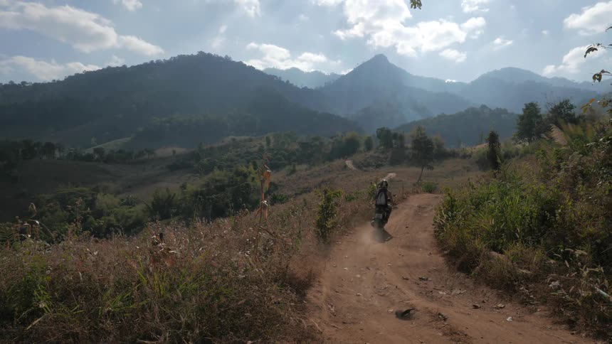 Man and woman ride motor scooters on dirt road in the field with mountains in the background