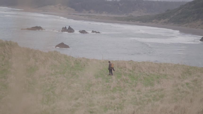 A lone figure walks across a grassy bluff overlooking a rugged coastline with sea stacks and crashing waves under an overcast sky