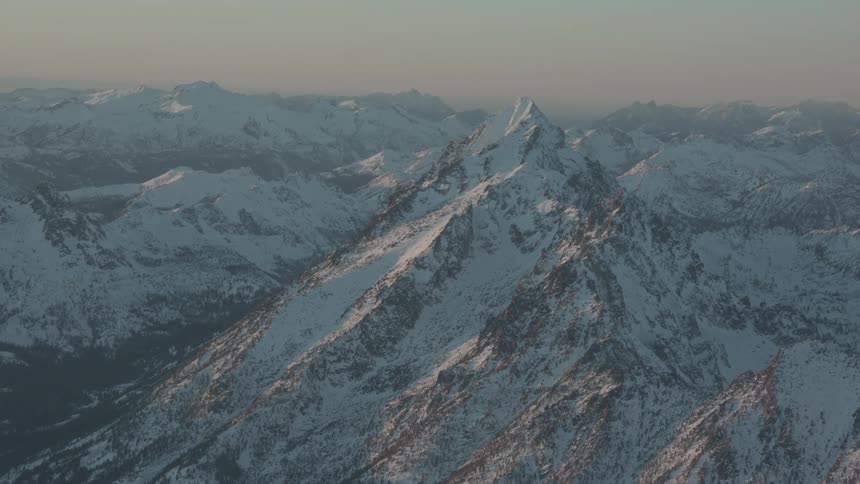 A majestic mountain peak covered in snow and surrounded by a vast mountain range under a soft hazy sky