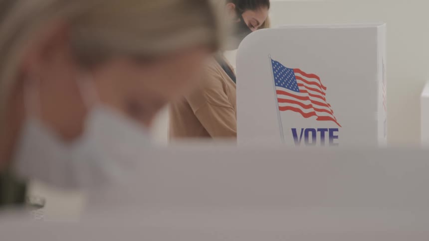 Two female american citizens wearing masks are casting their votes in a voting booth displaying the american flag and the word vote