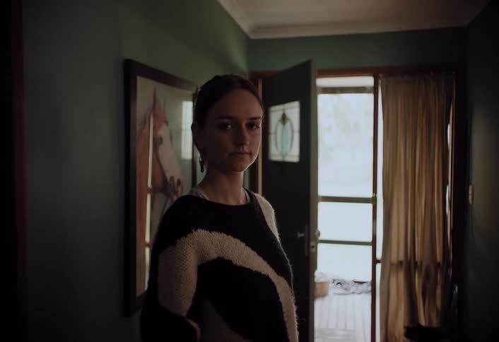 A woman with light hair and a striped sweater stands in a dimly lit hallway looking directly at the camera with a serious expression with an open doorway leading to a brighter room in the background