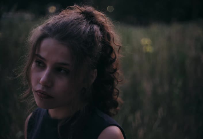 A young woman with curly brown hair pulled back in a loose bun looks pensively off camera with a soft natural background suggesting an outdoor setting at dusk