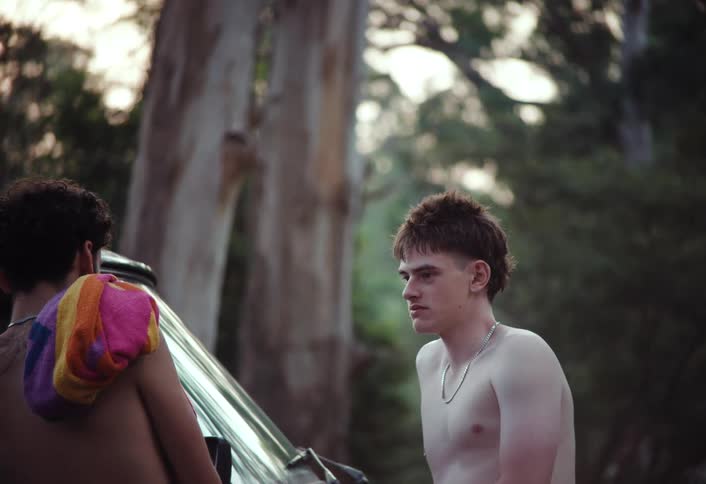 Two shirtless teenagers recline in a car talking with a forested area visible nearby